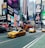 A bustling urban street scene with several iconic yellow taxis driving down a busy avenue. Bright, colorful advertisements and large digital billboards cover the facades of tall buildings. A double-decker tour bus and numerous pedestrians are visible, conveying a sense of vibrant city life.