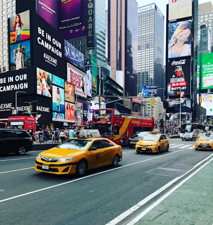 A bustling urban street scene with several iconic yellow taxis driving down a busy avenue. Bright, colorful advertisements and large digital billboards cover the facades of tall buildings. A double-decker tour bus and numerous pedestrians are visible, conveying a sense of vibrant city life.