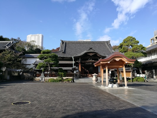 An ornate Buddhist temple with traditional architecture is set against a clear blue sky. The temple features a dark-tiled roof and ornate wooden carvings. In the foreground, an open courtyard is paved with gray stones, and a small wooden structure with a curved roof is visible.