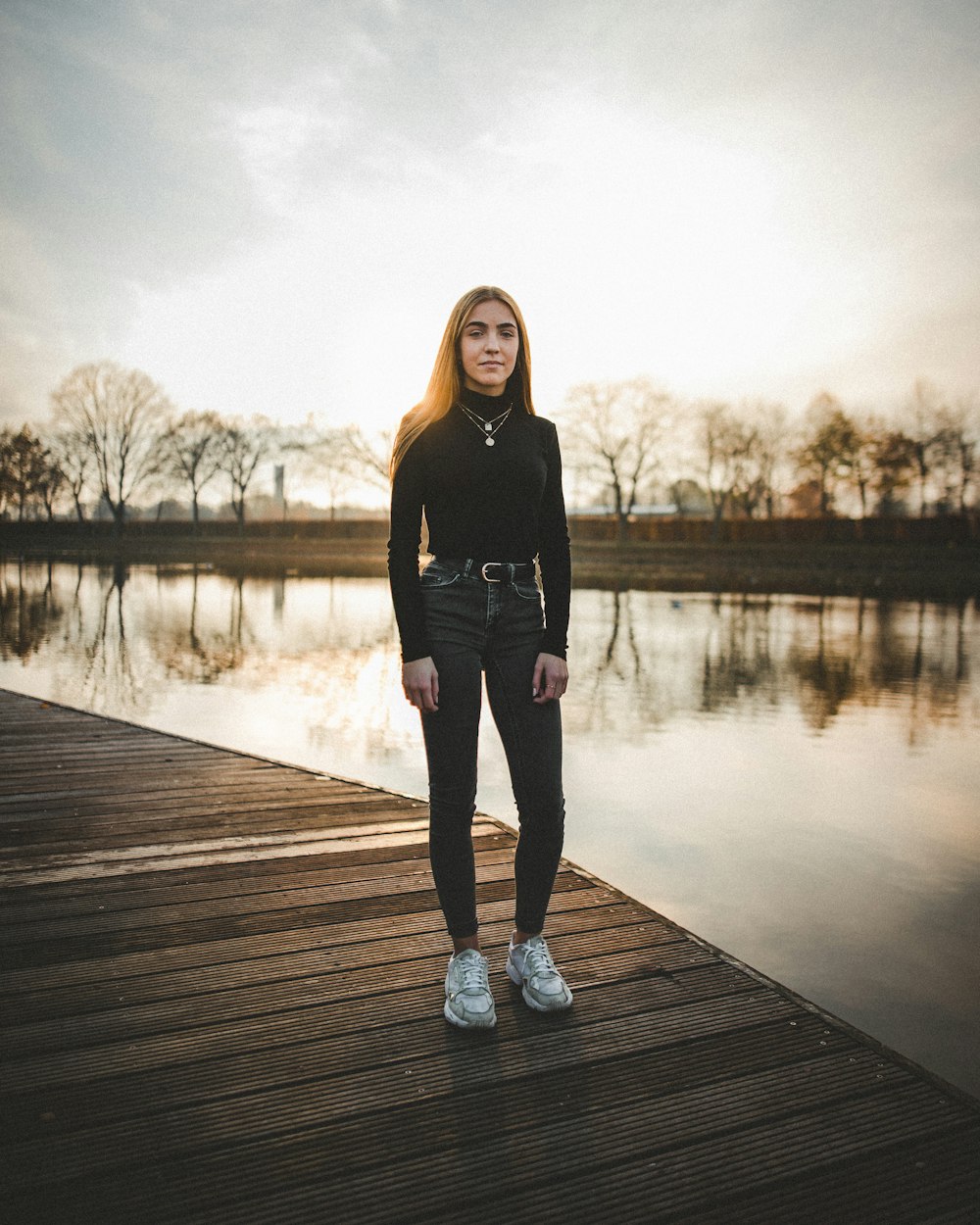 woman in black long-sleeved top and jeans standing on wooden dock during golden hour