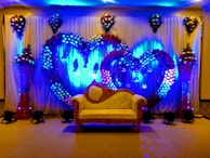A decorative event backdrop featuring two large intertwined heart shapes made of colorful flowers. The background is illuminated with vibrant blue lighting, enhancing the floral arrangement. There are cascading flower arrangements on tall stands placed symmetrically on either side, with additional floral clusters above. A vintage-style cream and brown sofa is placed at the center, adding to the elegance of the setting.