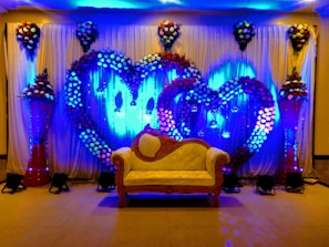 A decorative event backdrop featuring two large intertwined heart shapes made of colorful flowers. The background is illuminated with vibrant blue lighting, enhancing the floral arrangement. There are cascading flower arrangements on tall stands placed symmetrically on either side, with additional floral clusters above. A vintage-style cream and brown sofa is placed at the center, adding to the elegance of the setting.