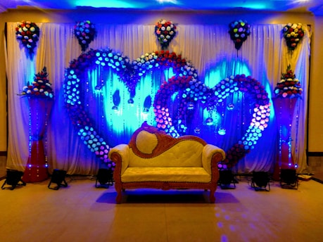A decorative event backdrop featuring two large intertwined heart shapes made of colorful flowers. The background is illuminated with vibrant blue lighting, enhancing the floral arrangement. There are cascading flower arrangements on tall stands placed symmetrically on either side, with additional floral clusters above. A vintage-style cream and brown sofa is placed at the center, adding to the elegance of the setting.