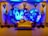 A decorative event backdrop featuring two large intertwined heart shapes made of colorful flowers. The background is illuminated with vibrant blue lighting, enhancing the floral arrangement. There are cascading flower arrangements on tall stands placed symmetrically on either side, with additional floral clusters above. A vintage-style cream and brown sofa is placed at the center, adding to the elegance of the setting.