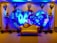 A decorative event backdrop featuring two large intertwined heart shapes made of colorful flowers. The background is illuminated with vibrant blue lighting, enhancing the floral arrangement. There are cascading flower arrangements on tall stands placed symmetrically on either side, with additional floral clusters above. A vintage-style cream and brown sofa is placed at the center, adding to the elegance of the setting.