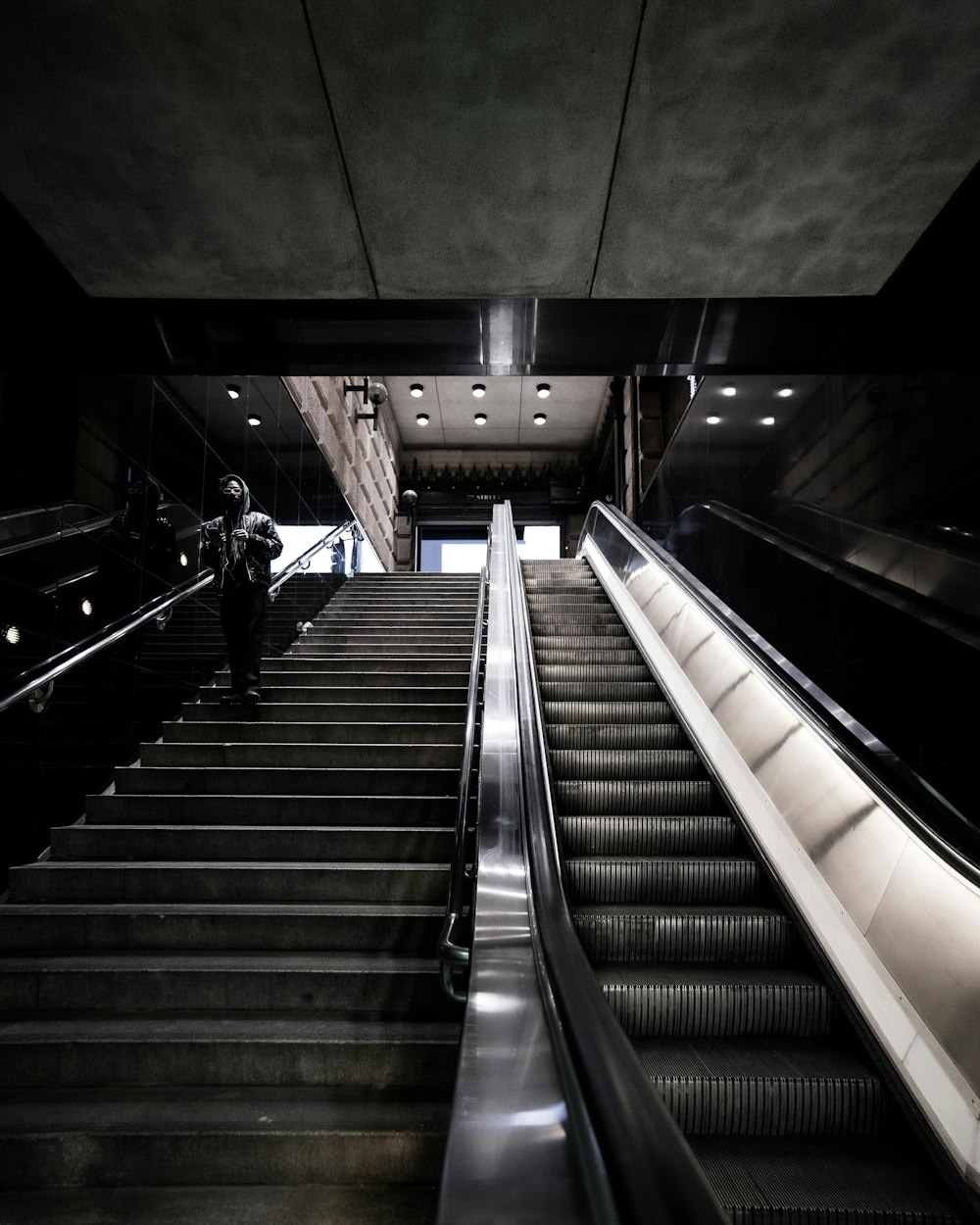 person wearing hooded jacket standing on concrete stairs near escalator