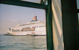 A large white cruise ship with colorful designs and the text 'Star Cruises' is docked in a harbor. The ship's name appears on the hull, and lifeboats are visible along its side. The image is taken from a building or another vessel, partially framed by a green-painted window structure, with calm water and a clear sky in the background.