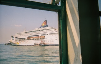 A large white cruise ship with colorful designs and the text 'Star Cruises' is docked in a harbor. The ship's name appears on the hull, and lifeboats are visible along its side. The image is taken from a building or another vessel, partially framed by a green-painted window structure, with calm water and a clear sky in the background.