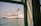 A large white cruise ship with colorful designs and the text 'Star Cruises' is docked in a harbor. The ship's name appears on the hull, and lifeboats are visible along its side. The image is taken from a building or another vessel, partially framed by a green-painted window structure, with calm water and a clear sky in the background.