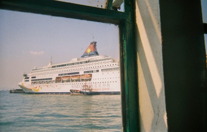 A large white cruise ship with colorful designs and the text 'Star Cruises' is docked in a harbor. The ship's name appears on the hull, and lifeboats are visible along its side. The image is taken from a building or another vessel, partially framed by a green-painted window structure, with calm water and a clear sky in the background.