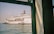 A large white cruise ship with colorful designs and the text 'Star Cruises' is docked in a harbor. The ship's name appears on the hull, and lifeboats are visible along its side. The image is taken from a building or another vessel, partially framed by a green-painted window structure, with calm water and a clear sky in the background.