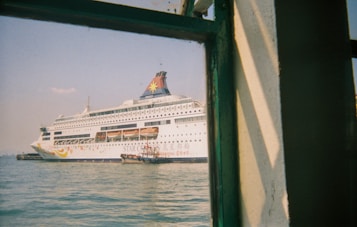 A large white cruise ship with colorful designs and the text 'Star Cruises' is docked in a harbor. The ship's name appears on the hull, and lifeboats are visible along its side. The image is taken from a building or another vessel, partially framed by a green-painted window structure, with calm water and a clear sky in the background.