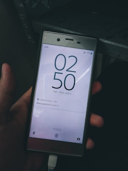 A hand holds a smartphone displaying a lock screen with the time 02:50 PM and the date Wednesday, January 8. The battery is at 77% and the phone is connected to a charger. A notification from the game 'Clash of Clans' is visible, mentioning 'Mine upgraded to level 3.' The phone seems to be in dark surroundings, and the design indicates an older model.