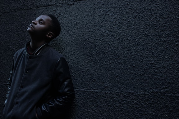 man wearing black button-up jacket standing and leaning against the wall while looking up