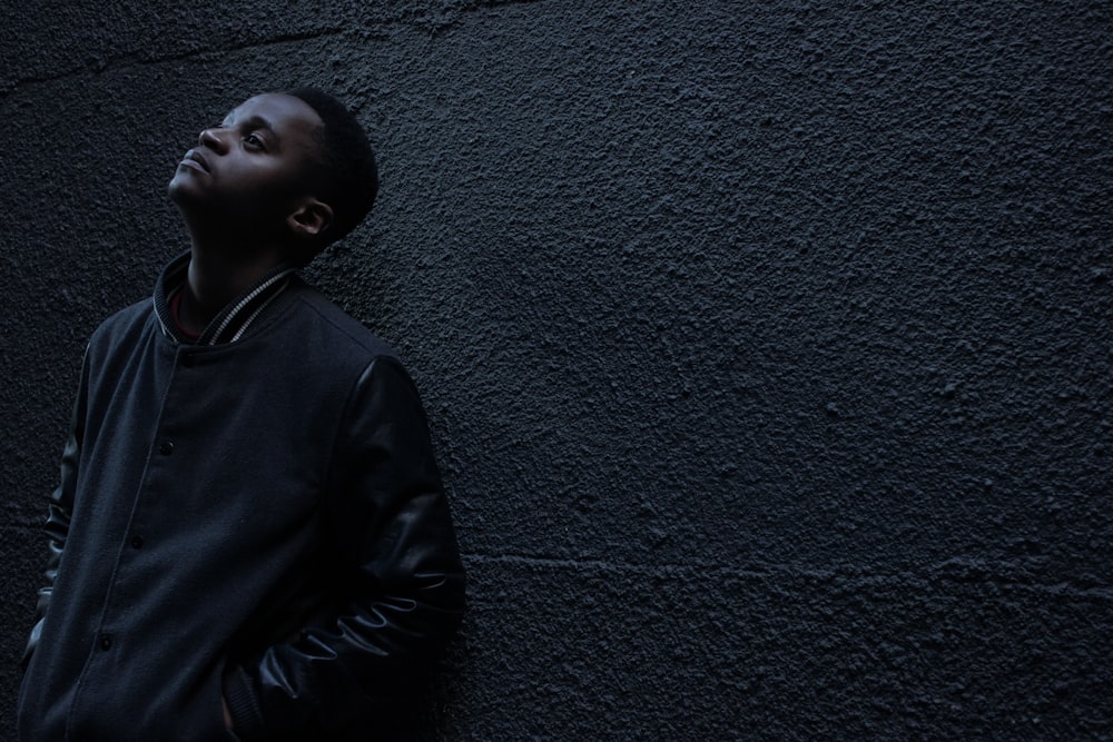 man wearing black button-up jacket standing and leaning against the wall while looking up