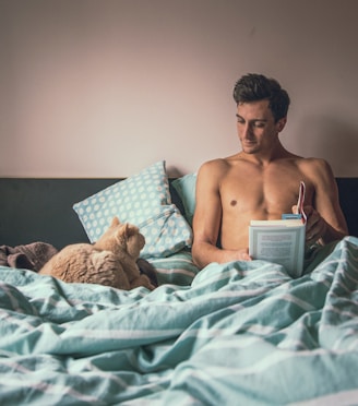 topless man reading book while sitting on bed near short-fur brown cat