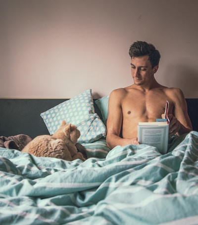 topless man reading book while sitting on bed near short-fur brown cat