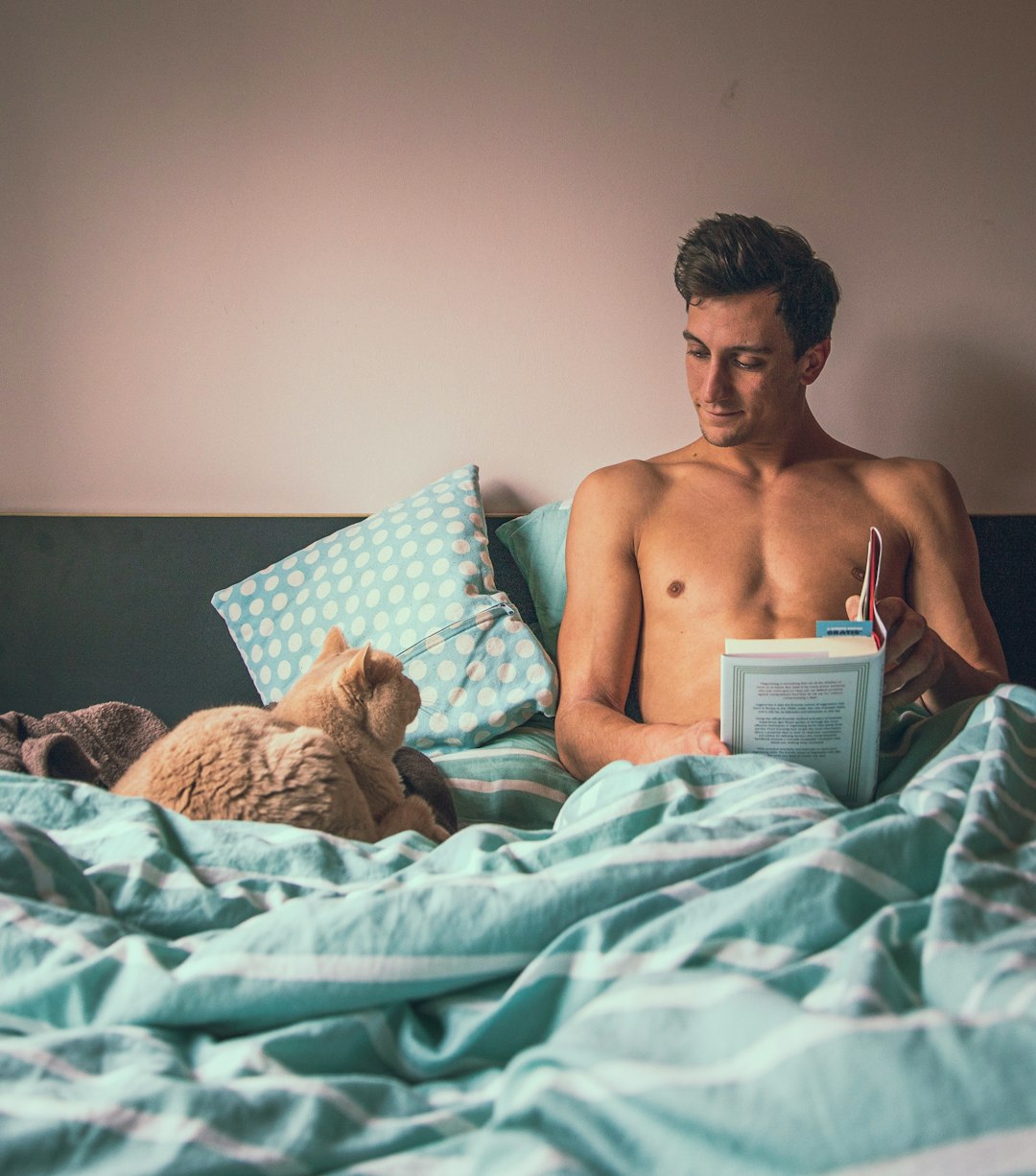 topless man reading book while sitting on bed near short-fur brown cat