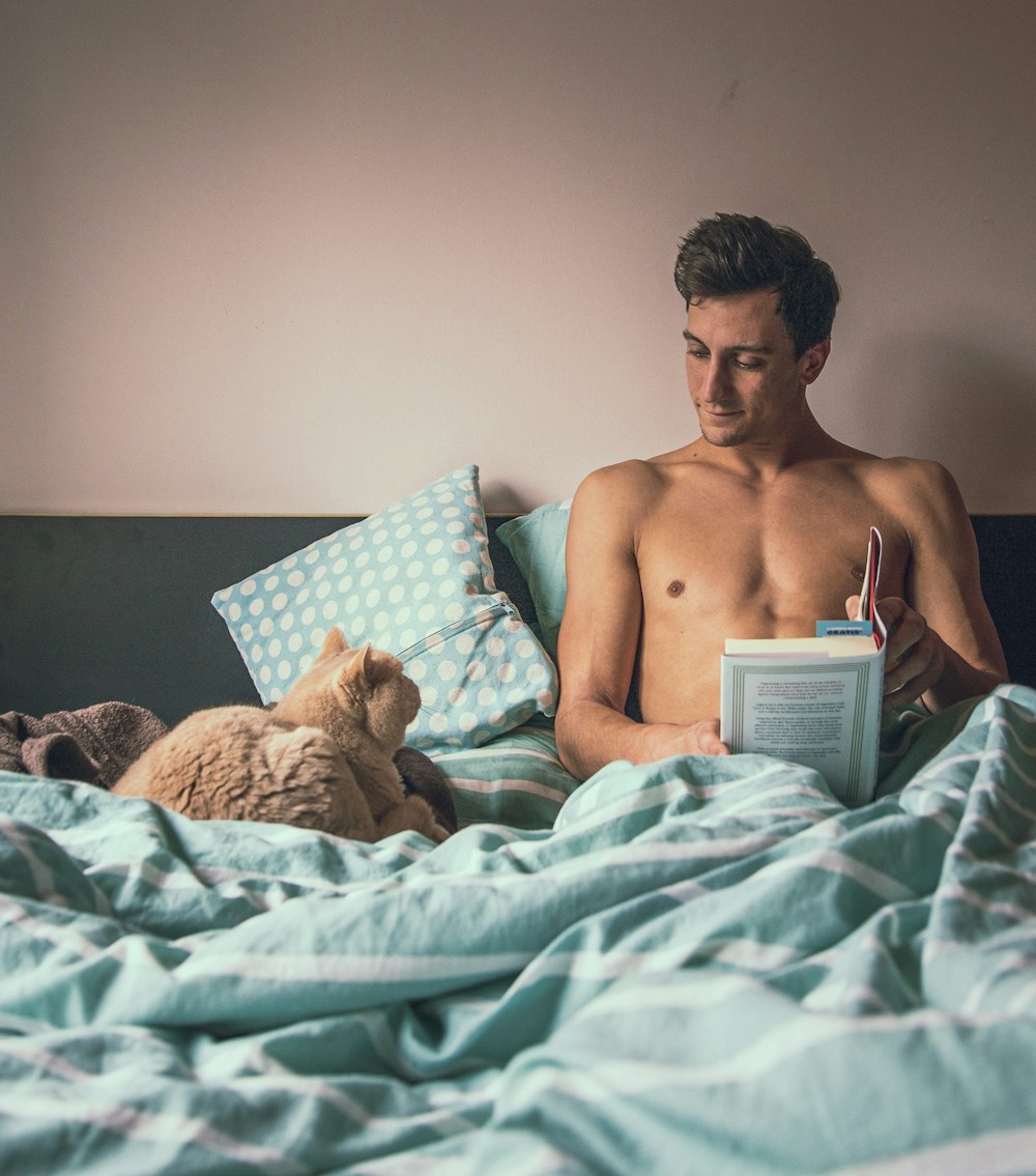 topless man reading book while sitting on bed near short-fur brown cat