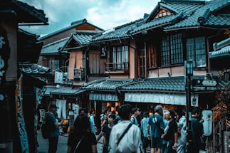 A bustling street scene featuring traditional Japanese architecture with wooden sliding windows and tiled roofs. A diverse group of people, including tourists and locals, are gathered, some walking, others conversing. Various signs with Japanese characters are visible, indicating shops or storefronts.