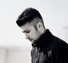 A monochromatic portrait of a person with well-groomed hair looking downward. The individual is wearing a black zip-up jacket, and the background is softly blurred, emphasizing the subject.