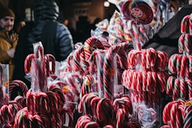 A variety of colorful candy canes are displayed for sale at a market stall, with several people browsing in the background.