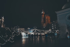 A nighttime scene featuring a large, ornate building with tall spires and a bustling marketplace. The area is adorned with festive string lights, creating a warm and inviting atmosphere. People are gathered around various stalls, adding a lively element to the scene.