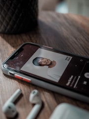 A mobile phone lies on a wooden surface, displaying an image of an album cover on a music app. Nearby, there are wireless earbuds and a case. The background has a textured pattern vase.