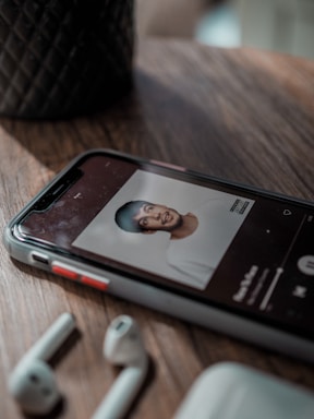 A mobile phone lies on a wooden surface, displaying an image of an album cover on a music app. Nearby, there are wireless earbuds and a case. The background has a textured pattern vase.