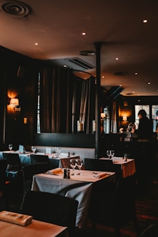 A dimly lit restaurant with neatly arranged tables covered in white tablecloths. Each table is set with wine glasses, cutlery, and napkins. Soft lighting from wall lamps and recessed ceiling lights creates a warm and cozy atmosphere. Curtains and a mirror are visible along the walls, enhancing the ambiance.