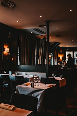 A dimly lit restaurant with neatly arranged tables covered in white tablecloths. Each table is set with wine glasses, cutlery, and napkins. Soft lighting from wall lamps and recessed ceiling lights creates a warm and cozy atmosphere. Curtains and a mirror are visible along the walls, enhancing the ambiance.