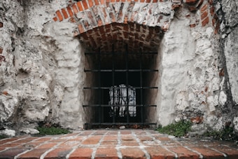 A small barred window set into a rough old stone wall, with bricks lining the arch above the opening. Some greenery is growing at the base.