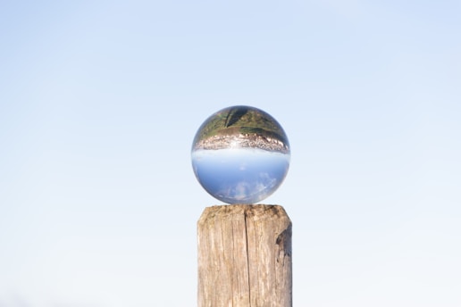 A crystal ball rests on the top of a wooden post. The ball reflects a landscape upside-down, showing a clear sky and greenery. The background is a pale blue sky, creating a serene atmosphere.