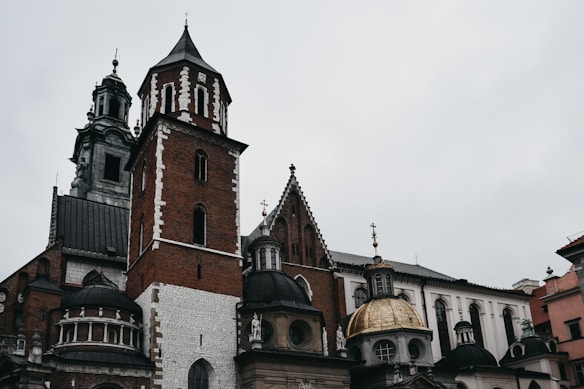 A historic architectural structure featuring multiple towers with intricate detailing. The buildings are constructed with red brick and white stone, featuring arched windows and domes. Some parts of the structure have ornate gold detailing.