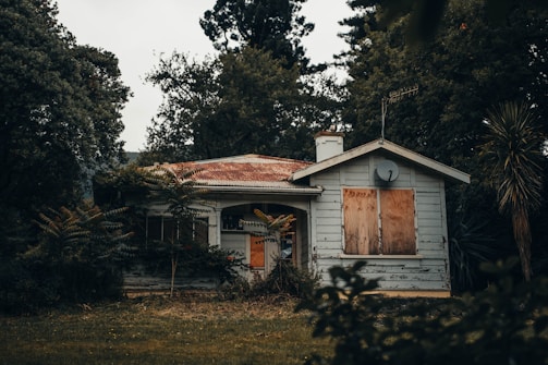 An old, abandoned house with boarded-up windows is surrounded by dense trees and foliage. The roof is rusted, and the paint on the house is peeling, indicating neglect. The yard is overgrown with grass and plants.