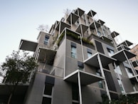 A modern, multi-story building features large, protruding balconies and an array of square windows. The design incorporates greenery, with plants and trees adorning various levels. The architecture is characterized by clean lines, geometric shapes, and a mix of neutral grey and black tones with patches of greenery.