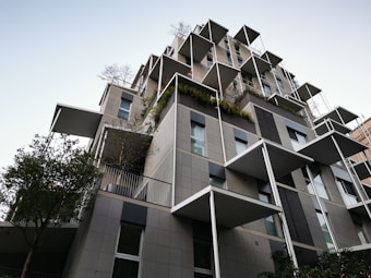 A modern, multi-story building features large, protruding balconies and an array of square windows. The design incorporates greenery, with plants and trees adorning various levels. The architecture is characterized by clean lines, geometric shapes, and a mix of neutral grey and black tones with patches of greenery.