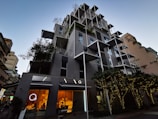 A modern, multi-story building with a creative architectural design featuring protruding square frames and greenery integrated into the structure. The lower floors have large glass windows with a warm glow from the lights inside, suggesting commercial or dining spaces. Trees adorned with string lights are positioned in front of the building, contributing to a sophisticated urban atmosphere.