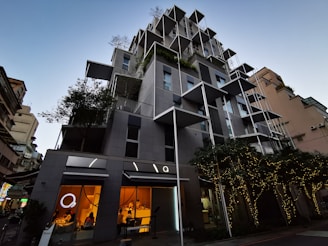 A modern, multi-story building with a creative architectural design featuring protruding square frames and greenery integrated into the structure. The lower floors have large glass windows with a warm glow from the lights inside, suggesting commercial or dining spaces. Trees adorned with string lights are positioned in front of the building, contributing to a sophisticated urban atmosphere.