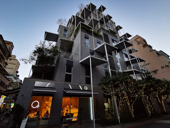 A modern, multi-story building with a creative architectural design featuring protruding square frames and greenery integrated into the structure. The lower floors have large glass windows with a warm glow from the lights inside, suggesting commercial or dining spaces. Trees adorned with string lights are positioned in front of the building, contributing to a sophisticated urban atmosphere.