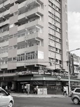 A multi-story residential building with visible wear and tear on its exterior. The street level features a bakery or café named 'Pastelaria Versalhes' with a green awning. People are walking on the sidewalk and cars are on the street.