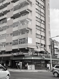 A multi-story residential building with visible wear and tear on its exterior. The street level features a bakery or café named 'Pastelaria Versalhes' with a green awning. People are walking on the sidewalk and cars are on the street.