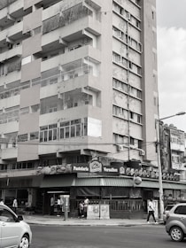 A multi-story residential building with visible wear and tear on its exterior. The street level features a bakery or café named 'Pastelaria Versalhes' with a green awning. People are walking on the sidewalk and cars are on the street.