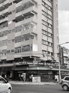 A multi-story residential building with visible wear and tear on its exterior. The street level features a bakery or café named 'Pastelaria Versalhes' with a green awning. People are walking on the sidewalk and cars are on the street.