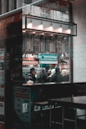 The scene captures a busy urban environment viewed through a window. A crowd of people is standing outside, some wearing winter clothing, suggesting cold weather. A sign for a currency exchange is prominently displayed, highlighting international travel or commerce. The architectural details of the building are visible in the background, featuring ornate stonework. The reflection on the window adds layers to the composition.