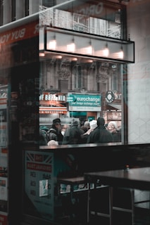 The scene captures a busy urban environment viewed through a window. A crowd of people is standing outside, some wearing winter clothing, suggesting cold weather. A sign for a currency exchange is prominently displayed, highlighting international travel or commerce. The architectural details of the building are visible in the background, featuring ornate stonework. The reflection on the window adds layers to the composition.