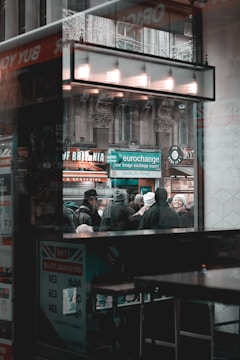 The scene captures a busy urban environment viewed through a window. A crowd of people is standing outside, some wearing winter clothing, suggesting cold weather. A sign for a currency exchange is prominently displayed, highlighting international travel or commerce. The architectural details of the building are visible in the background, featuring ornate stonework. The reflection on the window adds layers to the composition.