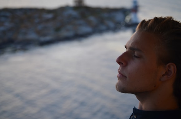A person with closed eyes and a calm expression is captured in profile, suggesting a moment of quiet reflection or meditation. The background is out of focus, showing a blurred body of water and possibly indistinct rocky formations or waves.