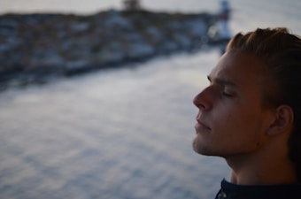 A person with closed eyes and a calm expression is captured in profile, suggesting a moment of quiet reflection or meditation. The background is out of focus, showing a blurred body of water and possibly indistinct rocky formations or waves.