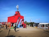 A vibrant red building with a rooftop antenna in a sandy area is surrounded by several people dressed in casual or sporty attire. There are large banners in front of the building, and a dining area with outdoor seating on the right. The sky is clear and blue, indicating a sunny day.